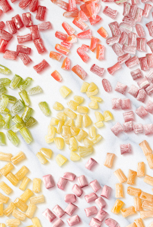 Assorted Butterfields Candy pieces in various flavors, including Peach, Cinnamon, Ginger, Key Lime, Lemon, Honeybell Orange, Cherry, Muscadine Grape, and Watermelon, arranged on a white marble surface. The candies have a glossy, sugar-dusted coating and come in vibrant colors, reflecting their unique flavors.